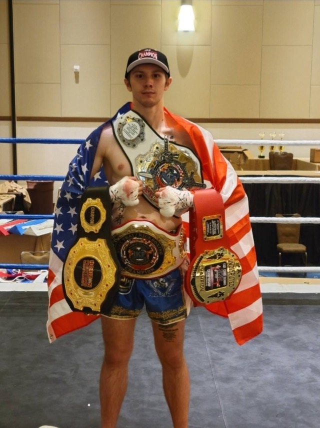 A kickboxer wearing several championship belts, draped in an American flag beside a ring