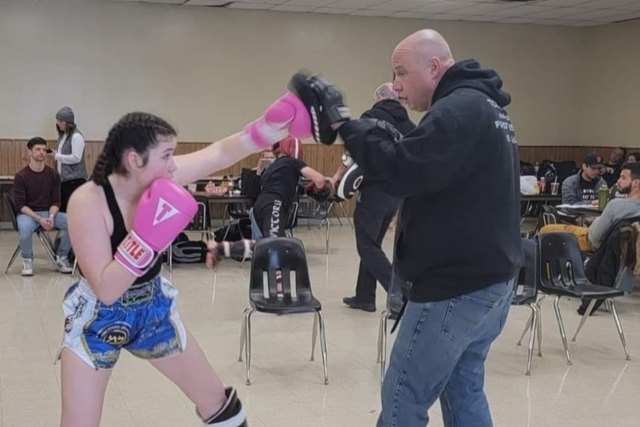 A coach holds focus mitts for a young kickboxer in pink gloves during class.