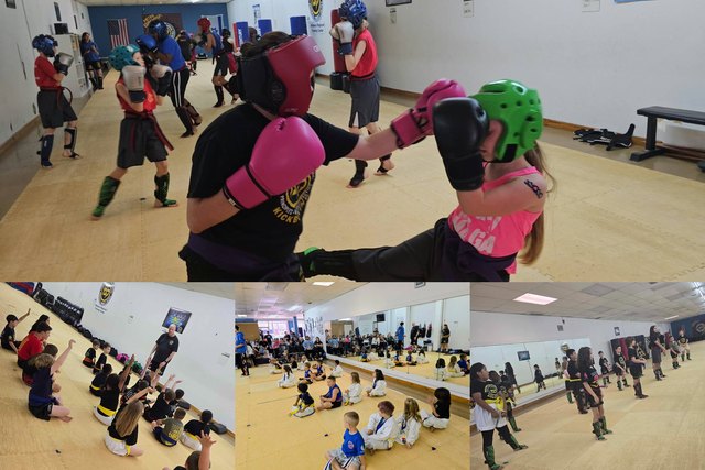 Two children in headgear and boxing gloves spar while classmates train behind them, with younger groups sitting on the mat listening to an instructor.
